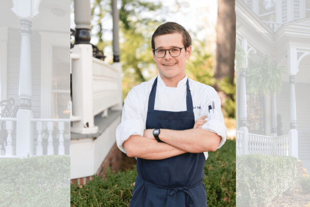 A smiling chef stands confidently with arms crossed in a charming outdoor setting.