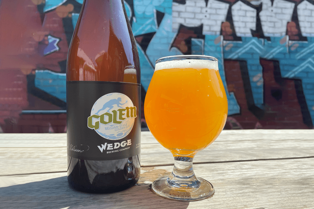 A bottle and glass of Golem Belgian-style golden ale sit on a wooden table against a colorful graffiti backdrop.