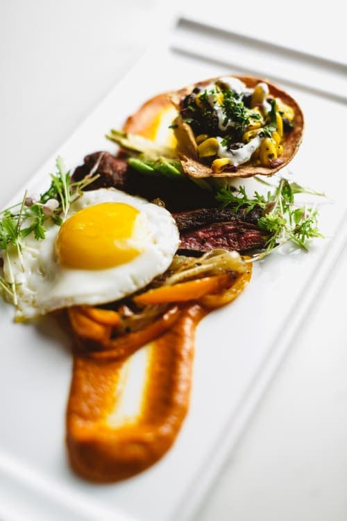 A gourmet plate featuring steak, a fried egg, roasted peppers, and a vegetable-filled tortilla cup.