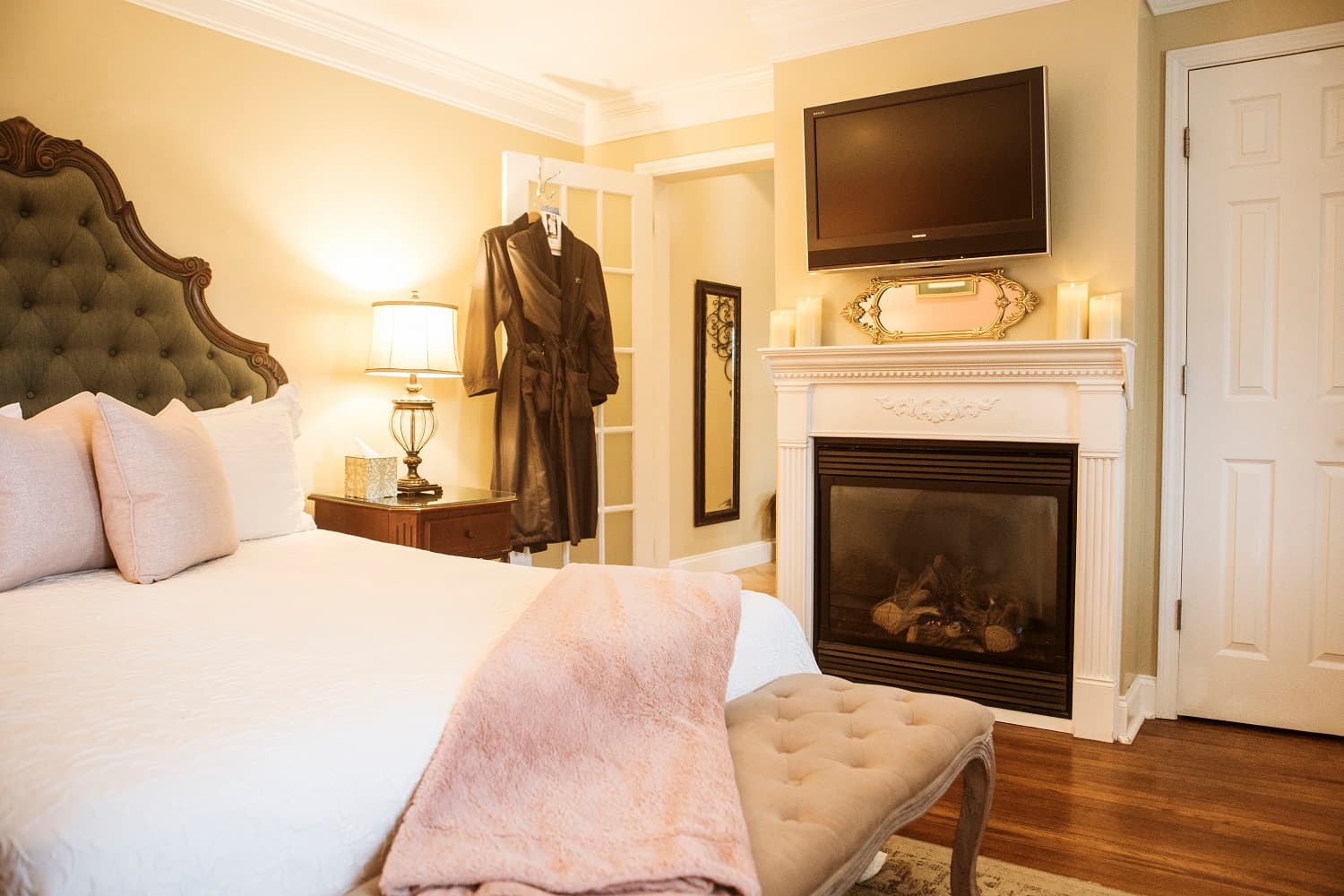 A cozy bedroom featuring a tufted headboard, soft pink pillows, a fireplace, and a flat-screen TV.