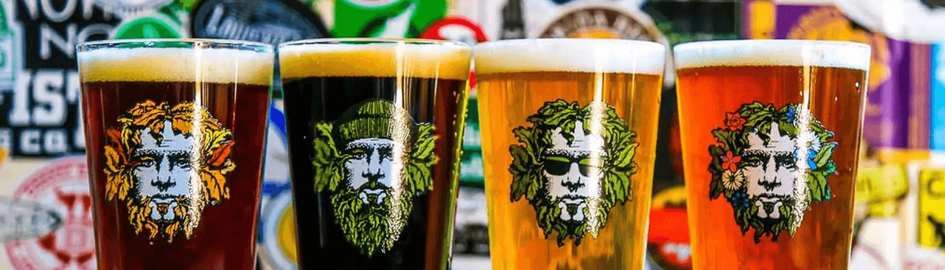 Four glasses of craft beer with colorful illustrated labels in front of a backdrop of brewery stickers.
