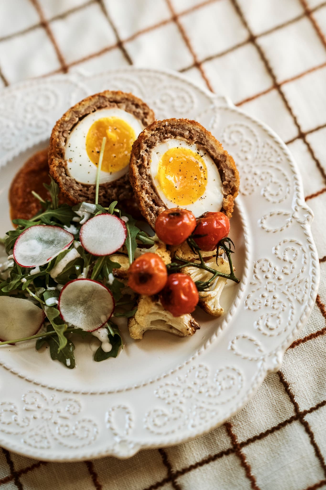 A plate of Scotch eggs, arugula salad, roasted cherry tomatoes, and sliced radishes.
