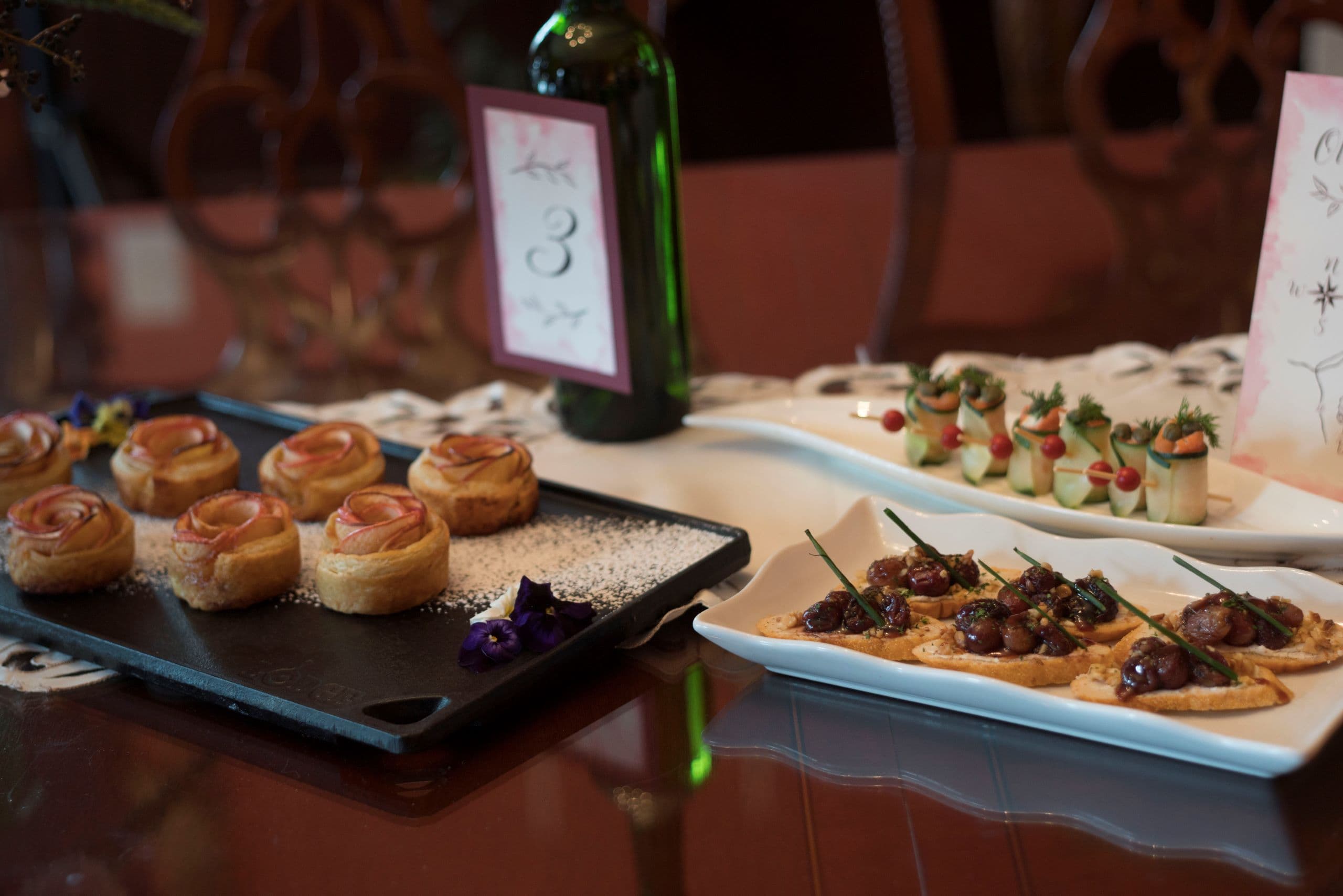 A table setting featuring elegant hors d'oeuvres including rose-shaped pastries, garnished bites, and a decorative wine bottle.
