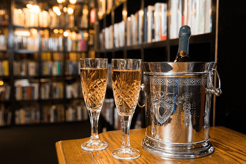 Two crystal glasses of sparkling rosé are placed next to a silver ice bucket containing a bottle of champagne, with bookshelves in the background.