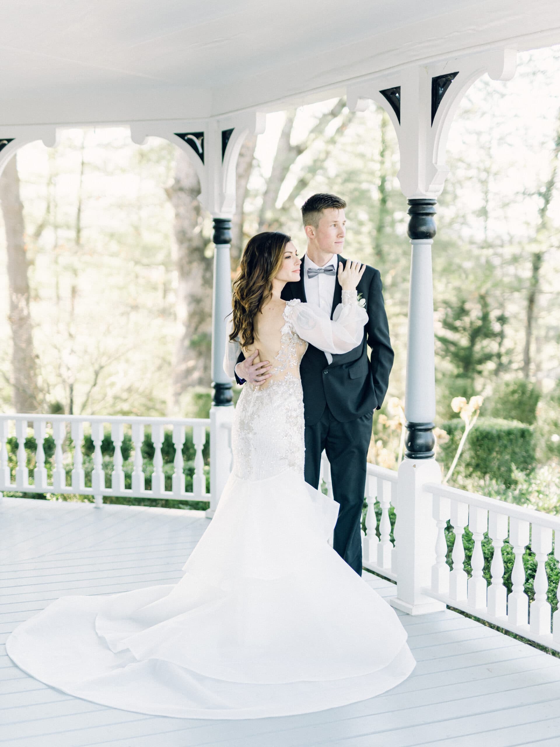 A couple in elegant formal attire poses romantically on a gazebo's porch, surrounded by lush greenery.